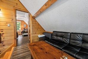 A cozy room with a wooden ceiling, a black leather sofa, a wooden table, and a bed with a quilt. - Lupine Lodge - Adorable, newly renovated cottage, with water access and dock on Loon Lake (Rangeley)