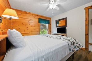 A bedroom with a bed, a TV, a ceiling fan, and a window with a view of greenery. - Lupine Lodge - Adorable, newly renovated cottage, with water access and dock on Loon Lake (Rangeley)