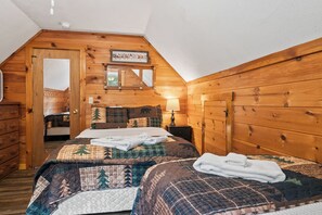 A cozy attic bedroom with a wooden sloped ceiling, a bed with a quilted bedspread, a nightstand with a lamp, and a mirror above the bed. - Lupine Lodge - Adorable, newly renovated cottage, with water access and dock on Loon Lake (Rangeley)