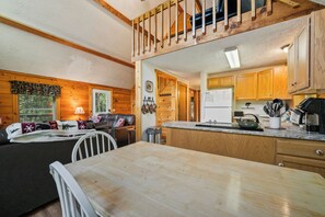 A cozy cabin interior with a dining table, a sofa, and a kitchen area with wooden cabinets and a refrigerator. - Lupine Lodge - Adorable, newly renovated cottage, with water access and dock on Loon Lake (Rangeley)