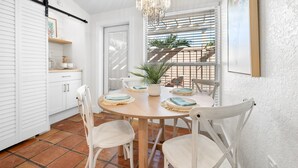 A dining area with a round wooden table, white chairs, a chandelier, and a view of a balcony with plants. - Plumeria Villa by Beach Boutique Rentals (Holmes Beach)