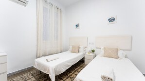 A hotel room with two beds, a window with sheer curtains, a small bedside table with a lamp and a plant, and a wall-mounted artwork.