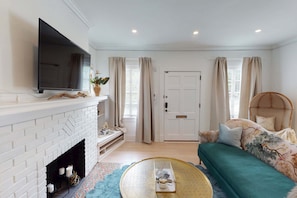 A living room with a white brick fireplace, a flat-screen TV mounted above it, a sofa with floral cushions, and a round coffee table. - Blithely Baroque (Atlanta)