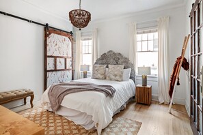 A bedroom with a bed, bedside lamps, a bench, a chandelier, and a large window with curtains. - Blithely Baroque (Atlanta)