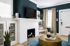 A living room with a white brick fireplace, a flat-screen TV mounted above it, a round gold coffee table, and a blue rug.