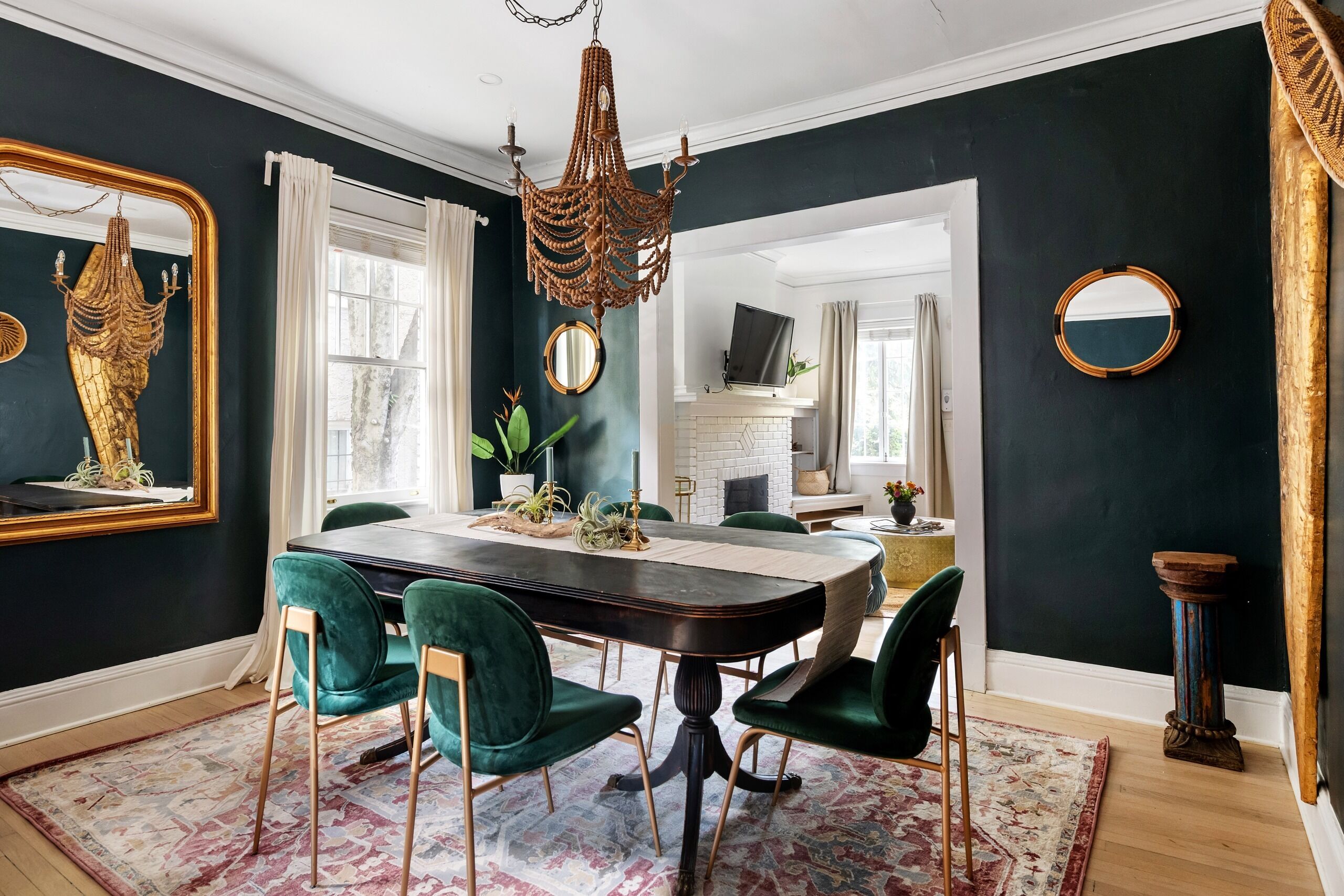 Un espace repas avec un mur de couleur sombre, un lustre, un grand miroir et des chaises recouvertes d’un tissu vert.