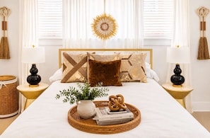 A neatly arranged bedroom with a large bed, decorative pillows, a bedside table with a lamp, a woven tray with books and a plant, and a sunburst wall decoration. - Blithely Baroque (Atlanta)
