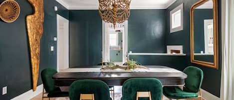 Comedor con pared oscura, lámpara de araña, sillas de terciopelo verde y un espejo grande.