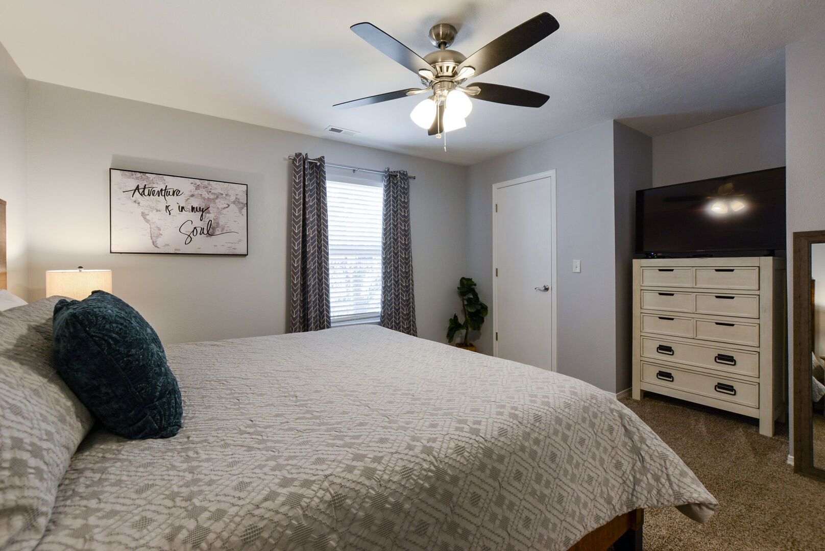 Une chambre à coucher comprenant un lit, un ventilateur de plafond, une commode, un téléviseur et un mur orné d’un cadre avec une citation.