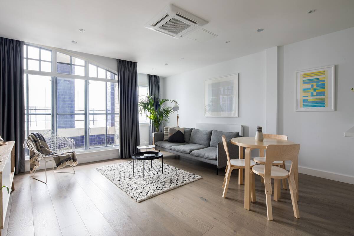 A modern living room with a sofa, a dining table with chairs, a white chair, a black coffee table, a patterned rug, and a large window with curtains.