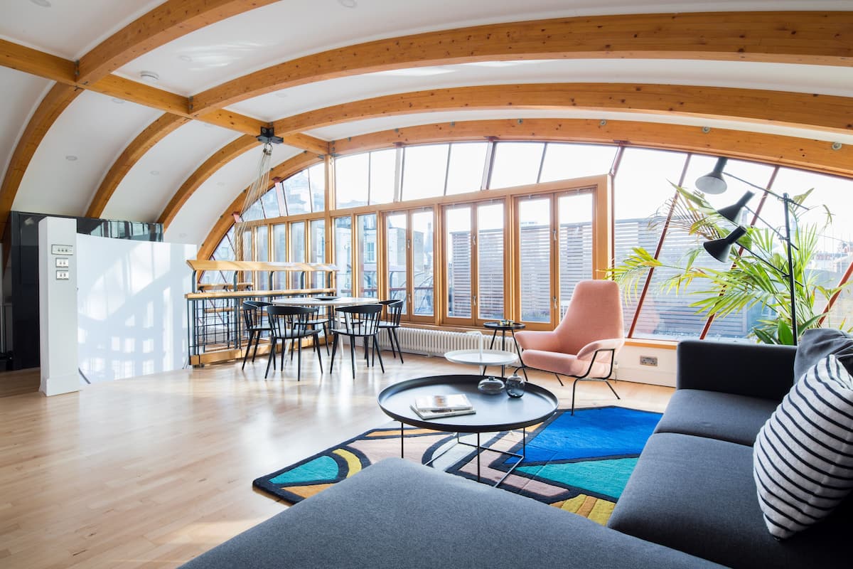 A spacious living area with a curved wooden ceiling, a dining area with a table and chairs, and a large window offering a view of the city.