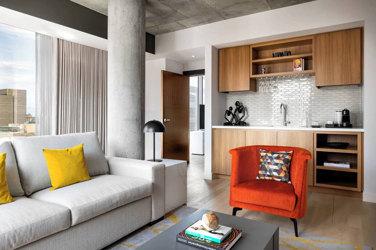 A modern living room with a sofa, a coffee table, and a red armchair. A kitchen area with wooden cabinets and a white tiled backsplash is visible in the background.
