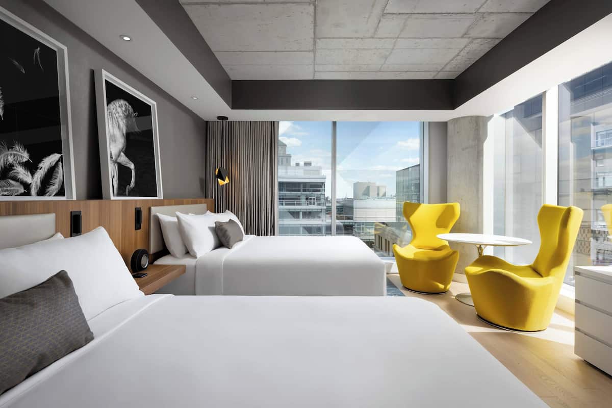 A modern hotel room with two beds, a large window with a city view, and yellow chairs.
