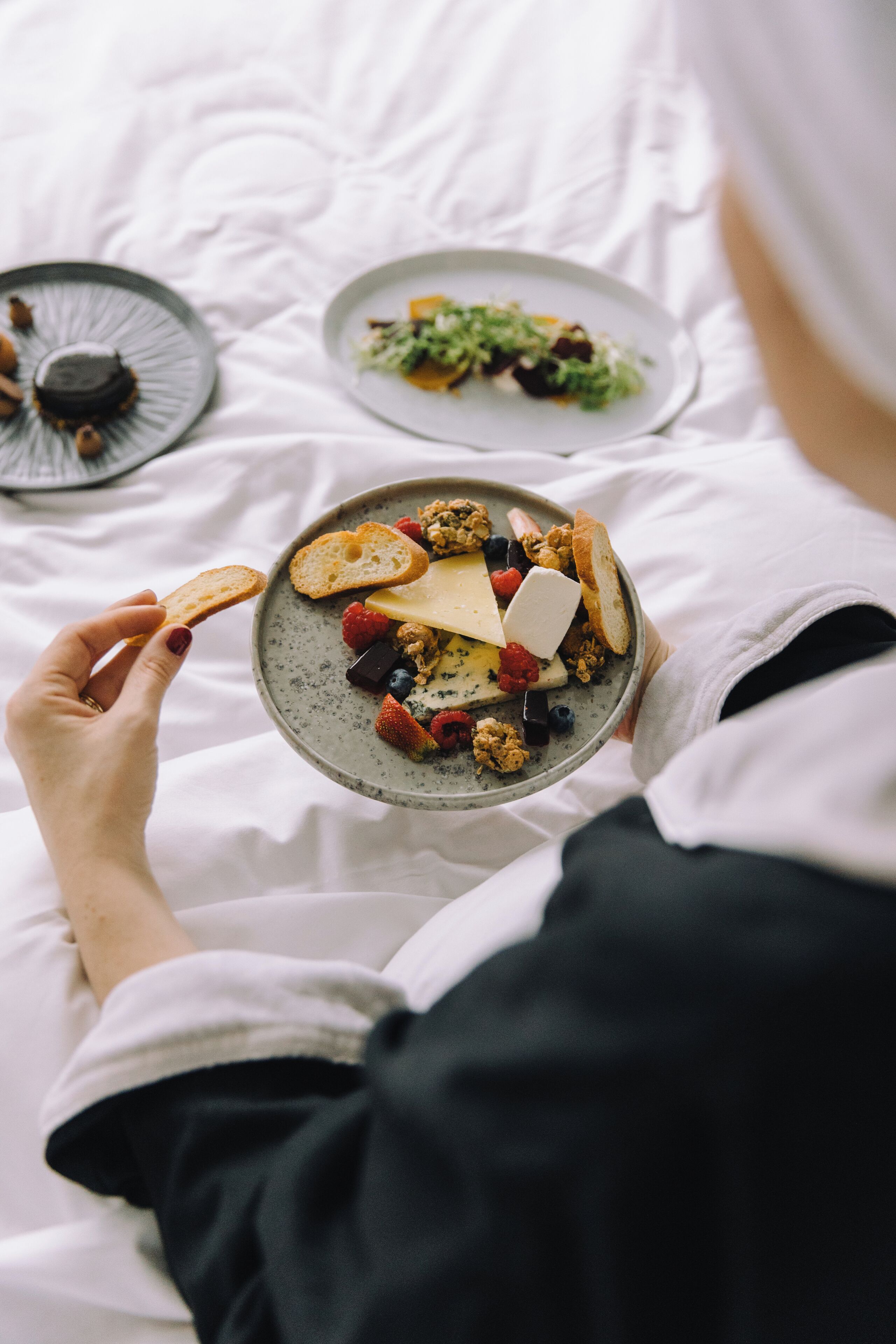 Une personne tenant une assiette remplie de divers aliments, notamment du fromage, du pain et des baies, sur un lit aux draps blancs.