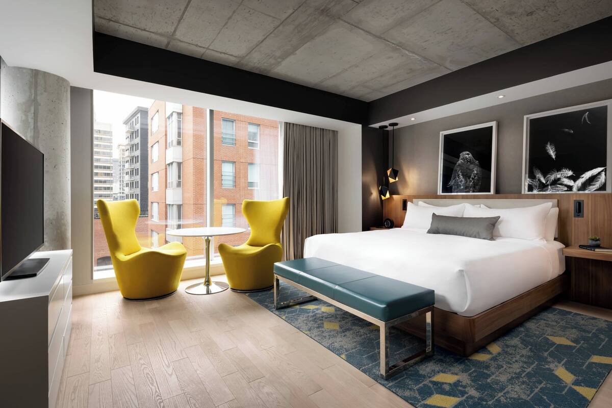 A modern hotel room with a large bed, a bench, two yellow chairs, a small table, and artwork on the walls.