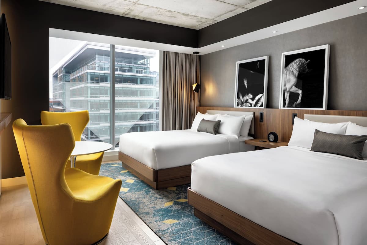 A hotel room with two beds, a yellow chair, a small table, and a large window with a city view.