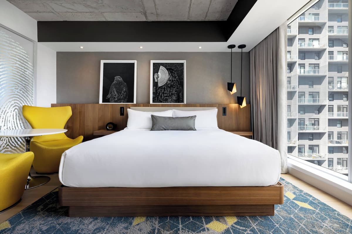 A modern hotel room with a large bed, yellow chairs, artwork on the wall, and a city view.