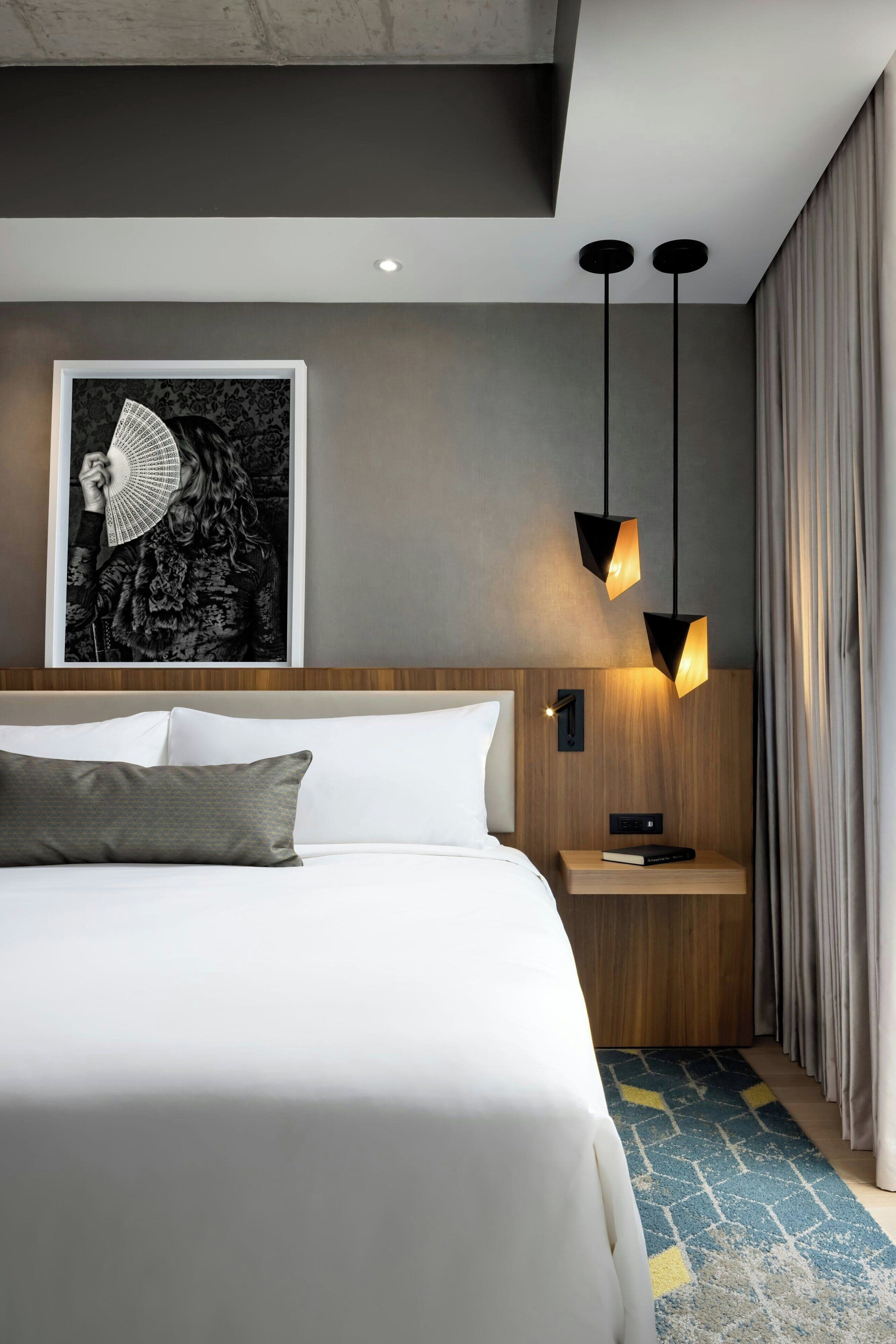 A modern hotel room with a large bed, a nightstand with a book, a framed picture on the wall, and two pendant lights.