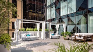 An outdoor seating area with round white sofas and a wooden bench, surrounded by greenery and a glass building facade. - Humaniti Hotel Montreal, Autograph Collection (Montreal)