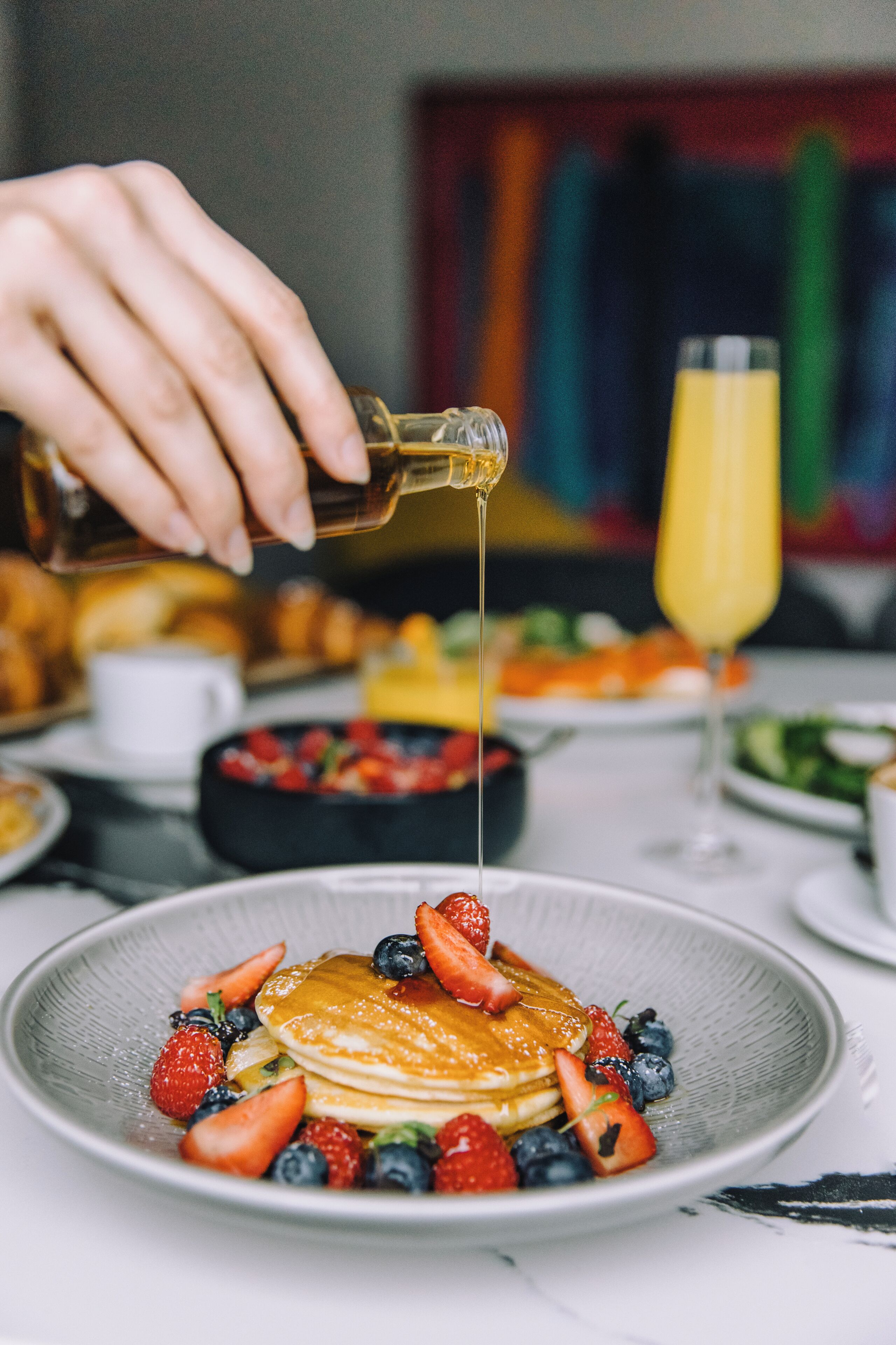 A breakfast setting with pancakes topped with strawberries and blueberries, syrup being poured, and a glass of orange juice in the background.