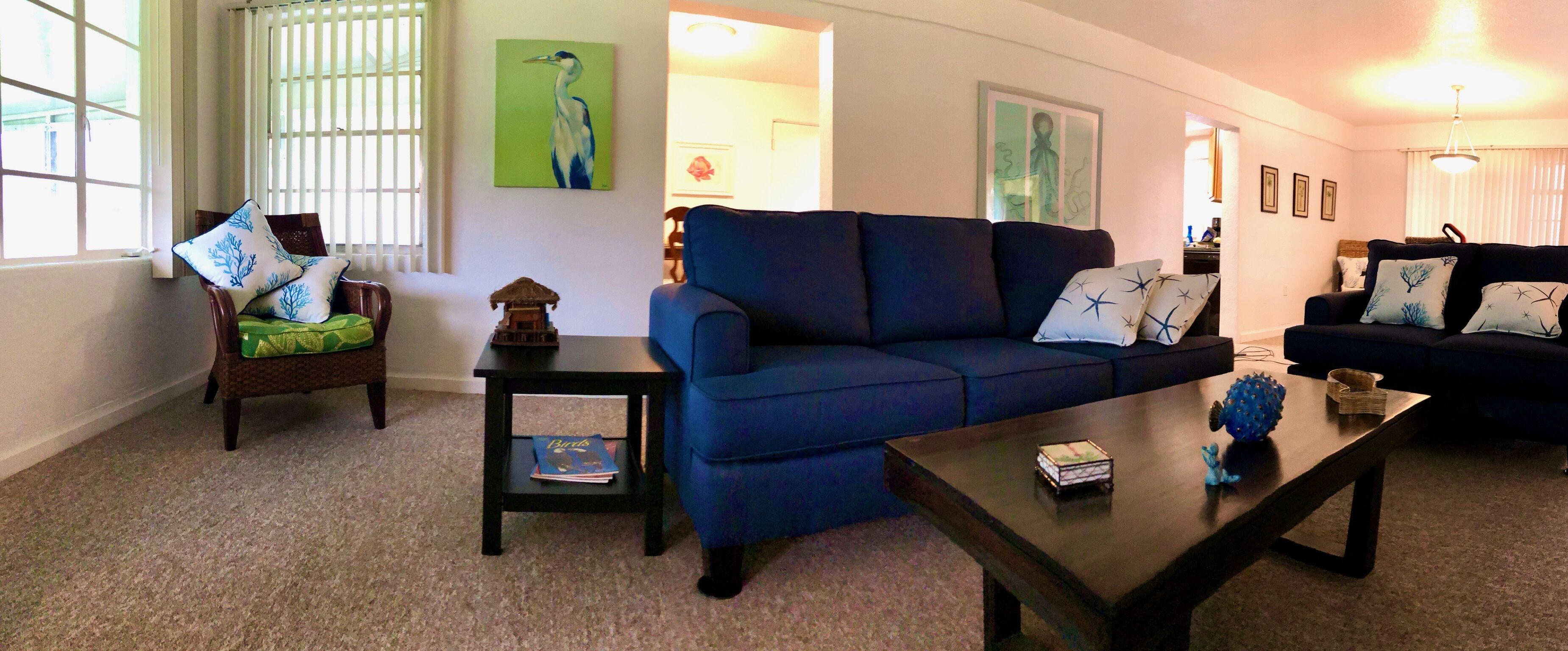 Una sala de estar con un sofá azul, una mesa de centro de madera y una silla con cojín. Hay cuadros enmarcados en la pared y una ventana con persianas.
