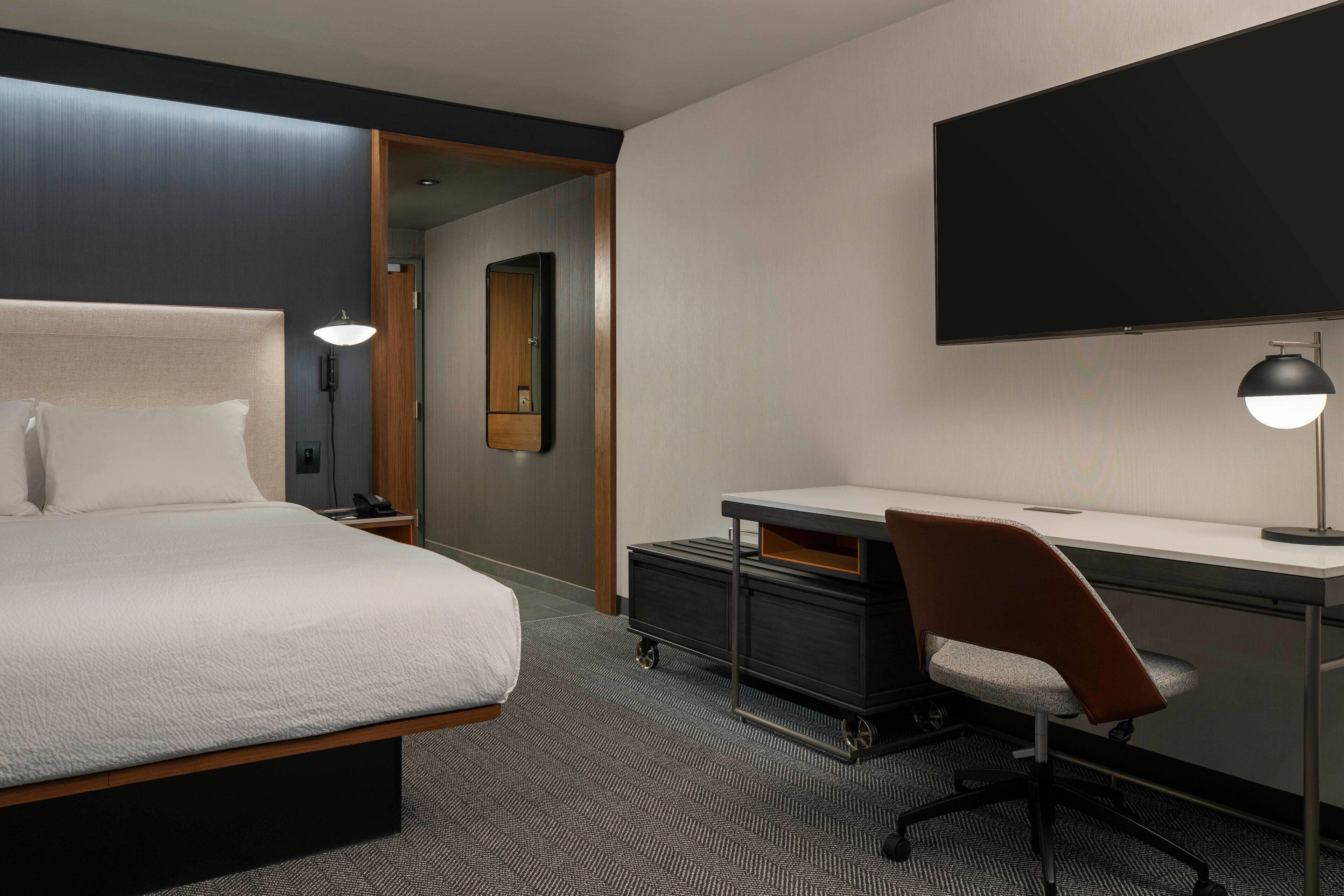A hotel room with a bed, desk, chair, and a flat-screen TV mounted on the wall.