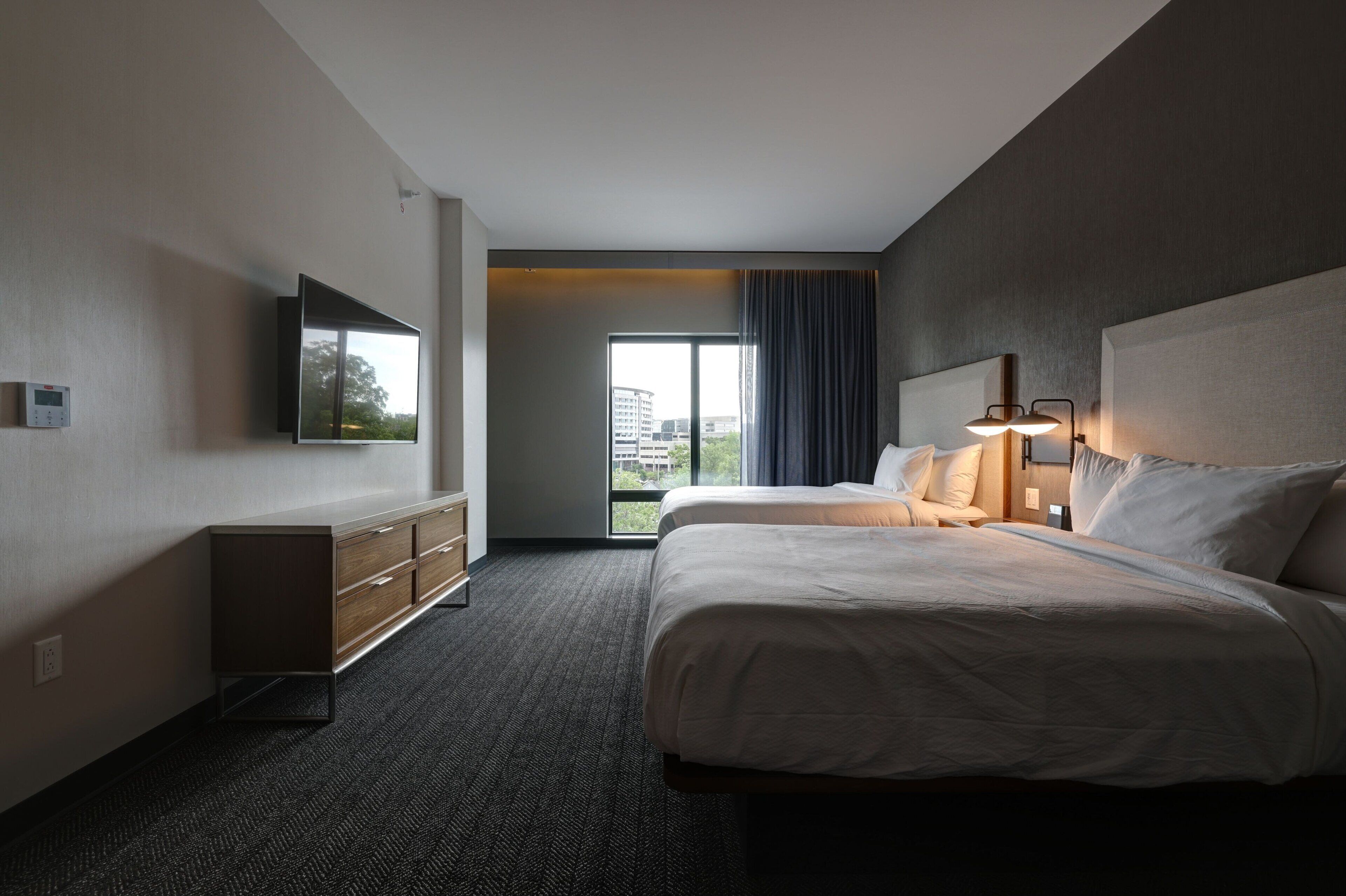 A hotel room with two beds, a flat-screen TV mounted on the wall, a dresser, and a large window with a view of buildings.