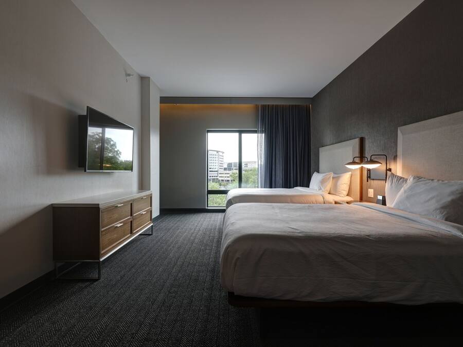 A hotel room with two beds, a flat-screen TV mounted on the wall, a dresser, and a large window with a view of buildings.
