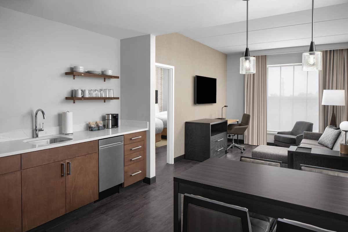 Una cocina moderna con armarios de madera, lavavajillas y una encimera con utensilios de cocina. Hay un comedor con mesa y sillas, y una sala de estar con sofá y televisor en la pared.