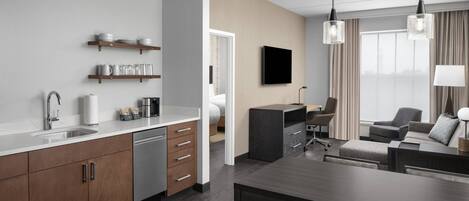 A modern kitchen with wooden cabinets, a dishwasher, and a countertop with kitchenware. There is a dining area with a table and chairs, and a living room with a sofa and a TV on the wall.