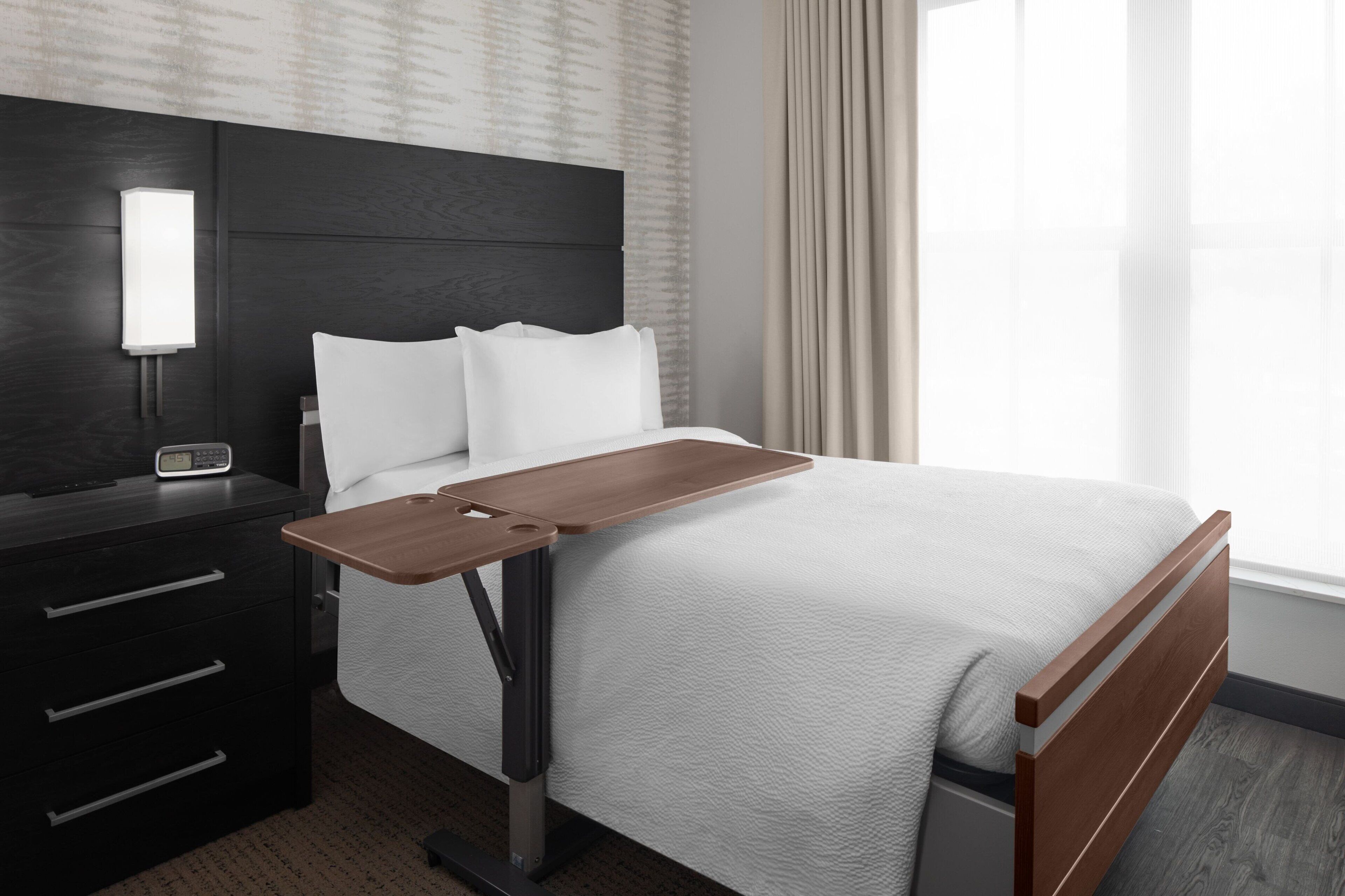 A modern hotel room with a bed, a bedside table with a lamp, and a window with curtains.