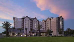 Un hotel de varios pisos con una entrada central, rodeado de césped cuidado y palmeras.