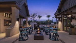 Un patio al aire libre con asientos y una chimenea central, rodeado de edificios y palmeras.