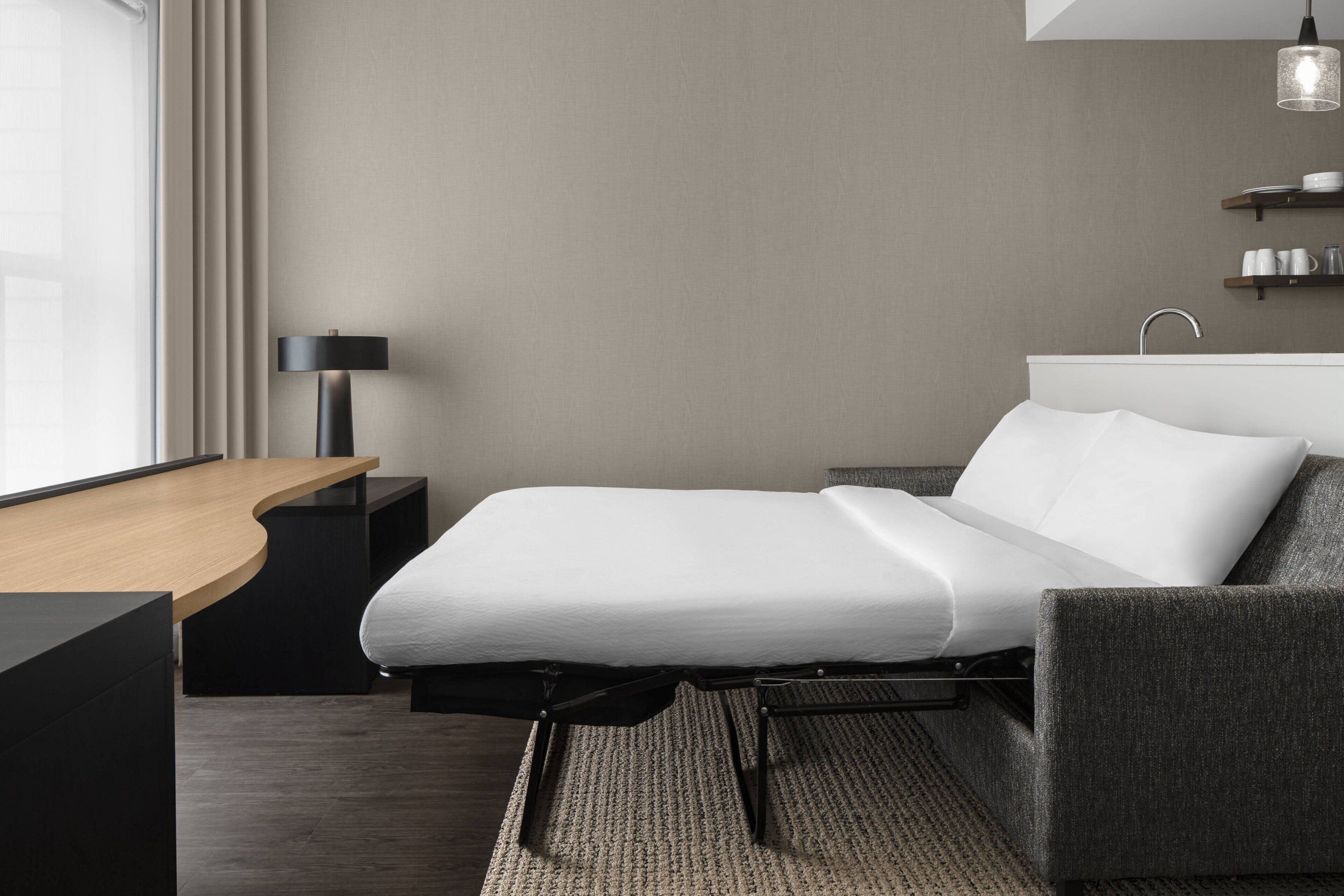 A modern hotel room with a bed, a desk, a lamp, and a view of the outdoors.