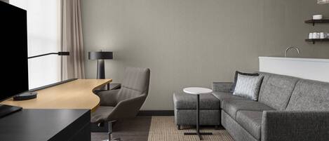 A modern hotel room with a desk, a grey sofa, a small round table, a chair, a TV, and a lamp.
