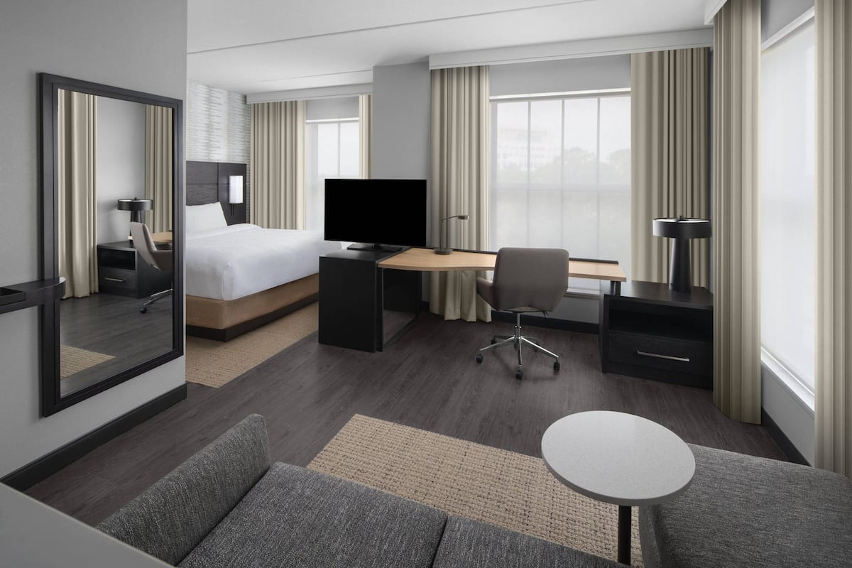 Habitación de hotel moderna con cama, escritorio, silla, televisor y ventanas grandes con cortinas.