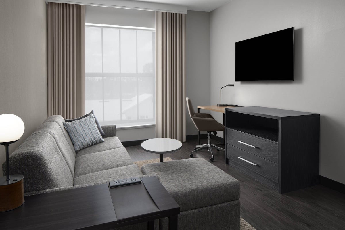 Una habitación de hotel moderna con un sofá gris, una mesita redonda pequeña, un escritorio con una silla y un televisor de pantalla plana montado en la pared.