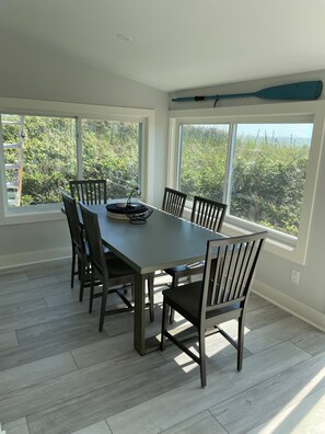 A dining area with a large table and chairs, large windows with a view of greenery, and a ceiling fan.