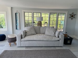 A living room with a beige sofa, a woven rug, a wicker side table, and a large window with a view of greenery.