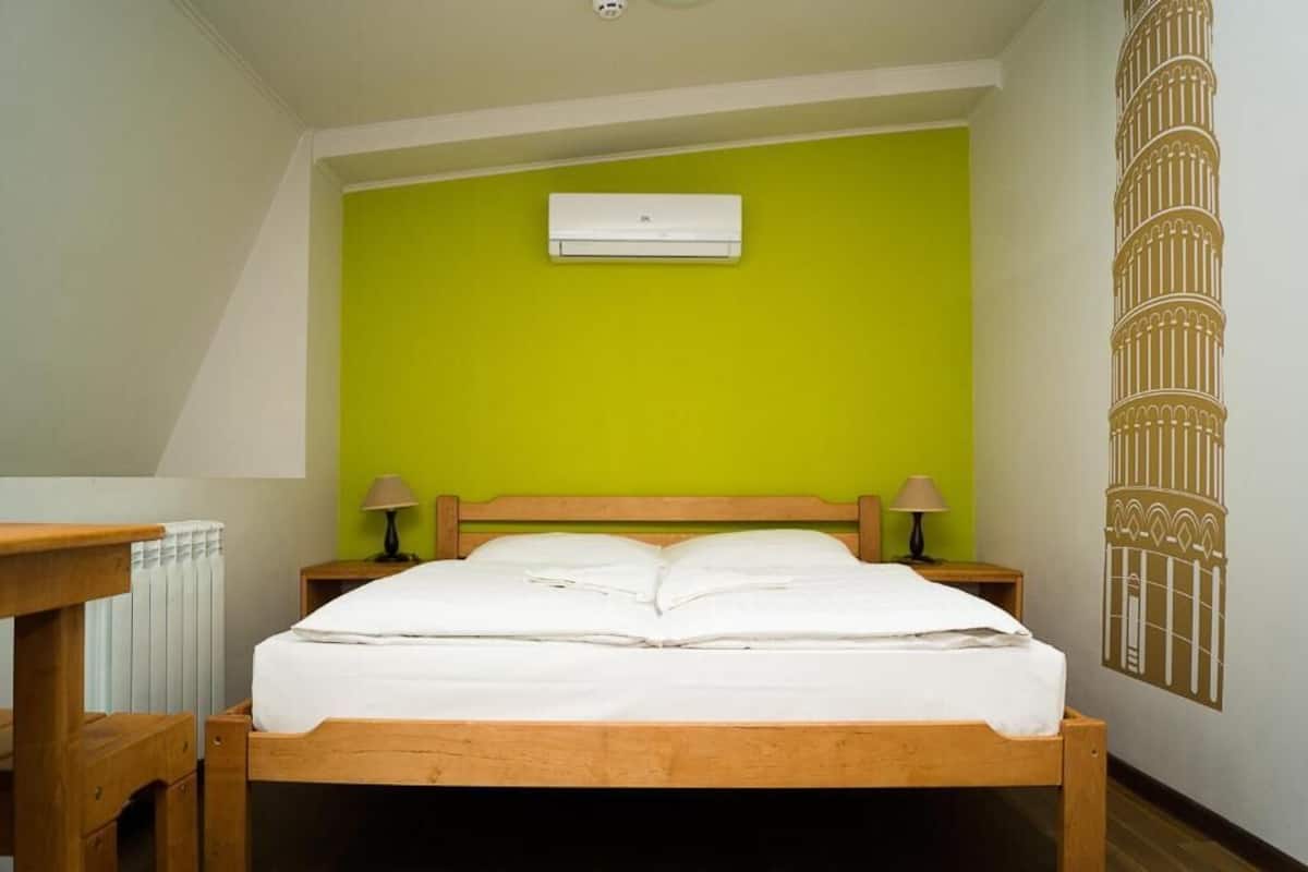 A bedroom with a green wall, a wooden bed frame, two bedside tables with lamps, and a white air conditioning unit on the wall.