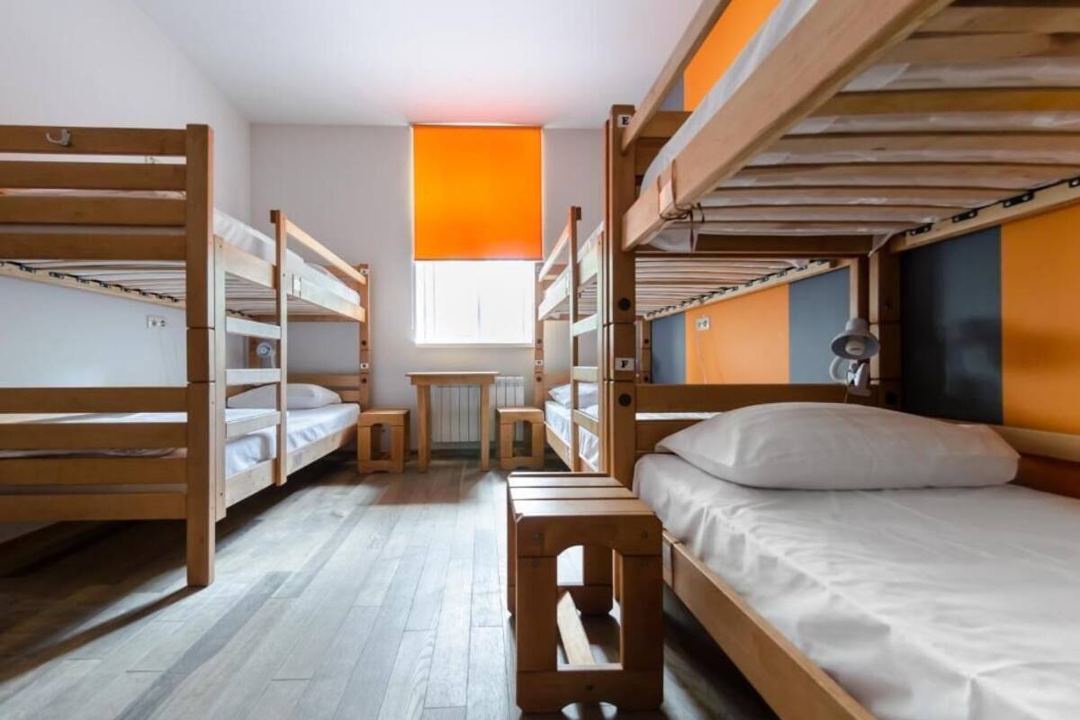 A dormitory room with bunk beds, a small table, and a window with an orange blind.
