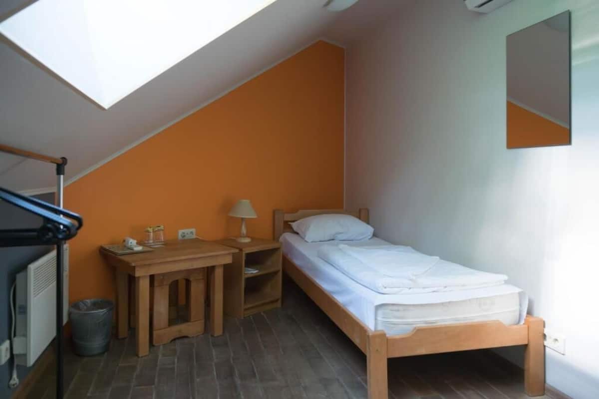 A small, single-bed room with a sloped ceiling, a wooden nightstand, a lamp, and a radiator.