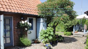 Een landelijk pand met een pannendak, een houten deur en een bakstenen muur. Er is een tuin met verschillende potplanten en een houten bank.