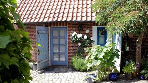 Een bakstenen huis met een pannendak, een blauwe deur en een tuin met potplanten en een boom.