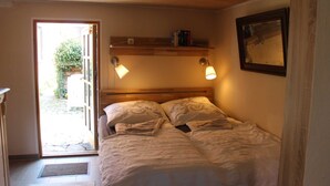 Een slaapkamer met een houten hoofdbord, twee bedlampen, een klein plankje met boeken en een deur naar een tuin.
