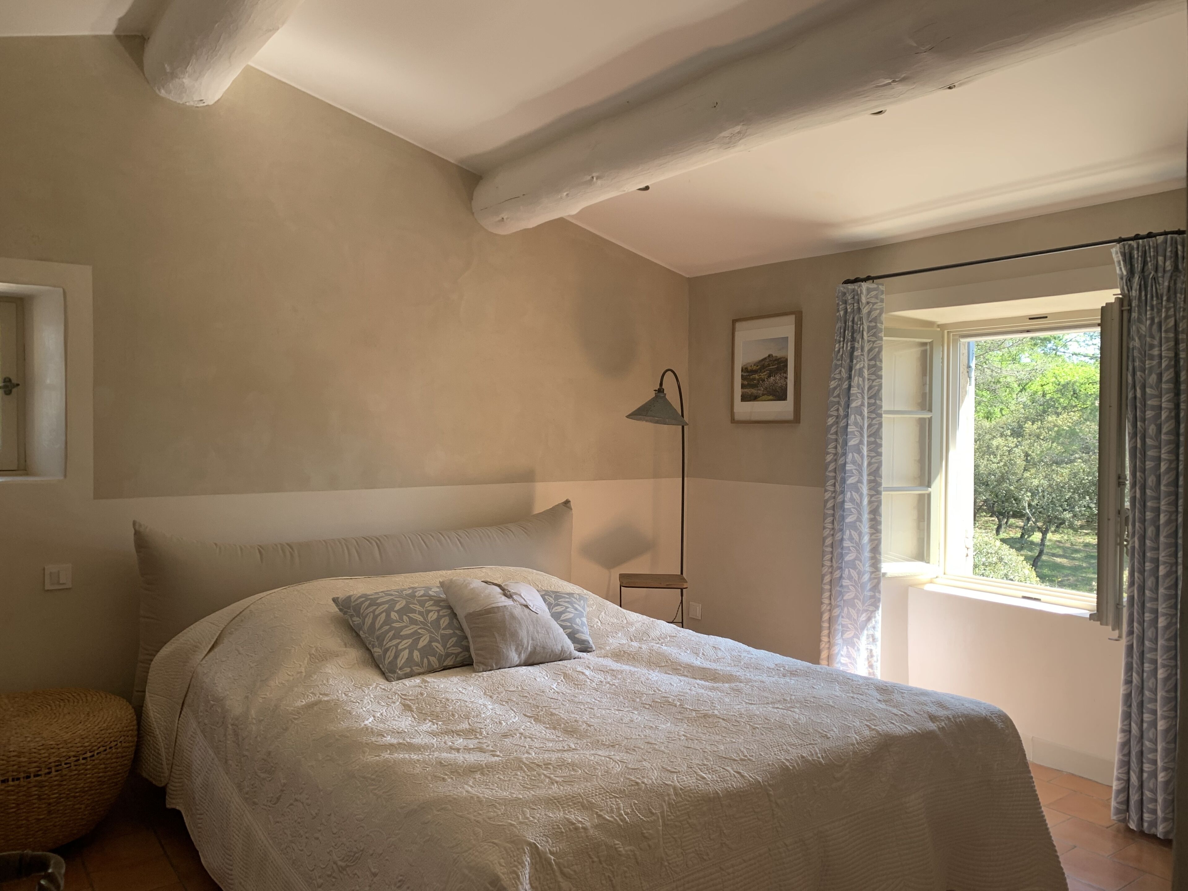 Un dormitori amb un llit gran, una làmpada de nit, una finestra amb cortines i un quadre al mur.