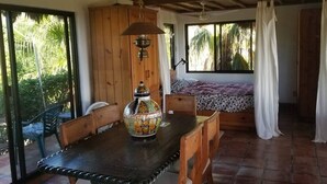 Una habitación con una mesa de madera, sillas, un jarrón decorativo, una cama con colcha estampada y una ventana con cortinas.