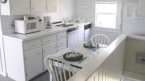 A compact kitchen with white cabinets, a microwave, a stove, and a sink. There is a dining area with a table and chairs. - The Whistling Whale - Fantastic Location!! (Chatham)