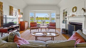 Un salon équipé d’une télévision, de deux fauteuils, d’un canapé, d’une cheminée et offrant une vue sur la verdure grâce à de grandes fenêtres.