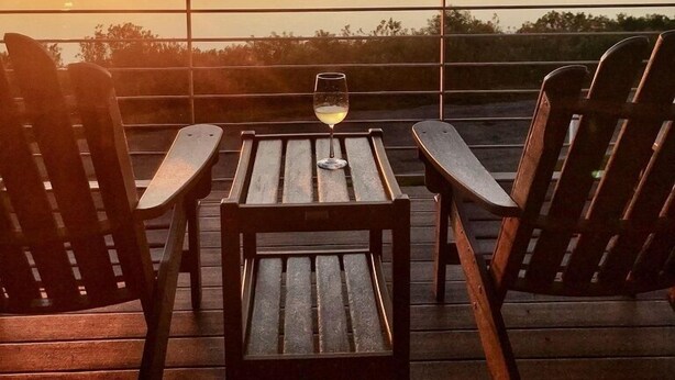 A sunset view from a deck with two Adirondack chairs and a table, overlooking a body of water.